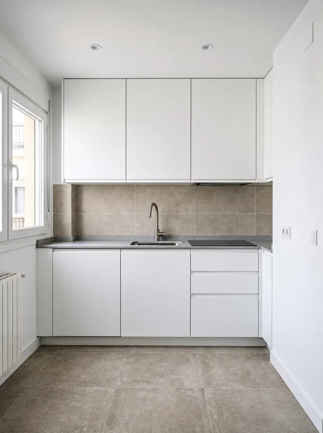 Cocina reformada con frentes blancos, encimera gris, campana integrada y ventana a fachada